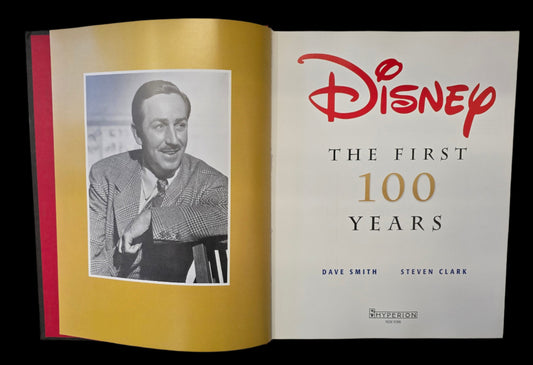 A book; Disney: The First 100 Years, opened to the title page, with a black and white photograph of Walt Disney on a gold colored page.