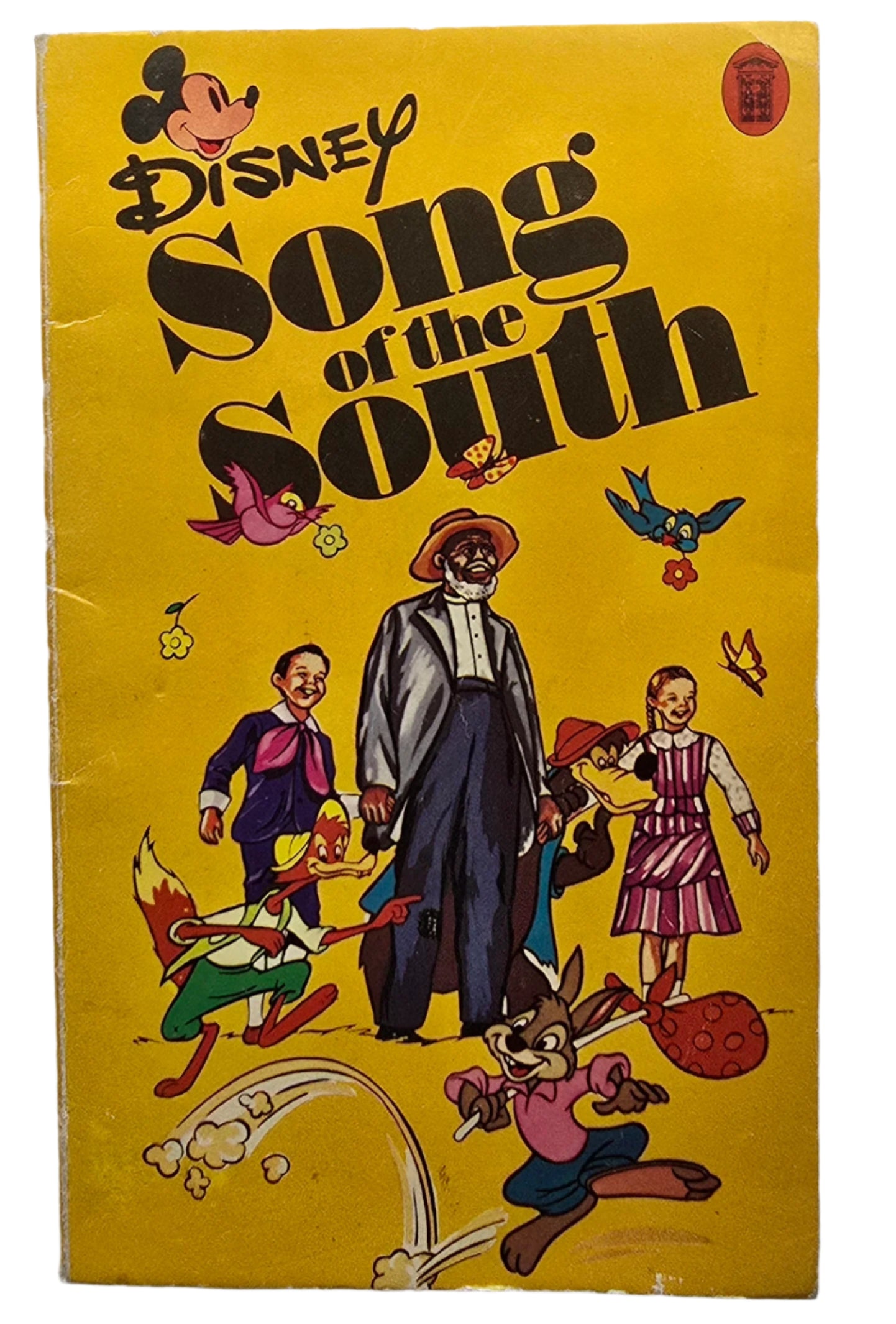 A copy of a paperback travel book; Disney's Song of the South from 1975, an adaptation by Guy N Smith. It features Uncle Remus, Br'er Rabbit, Br'er Fox, Br'er Bear, and Ginny and Jake on a yellow background. 